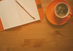 An open lined notebook on a wooden surface, with a pencil on top of it. To the right is an orange cup and saucer with coffee.