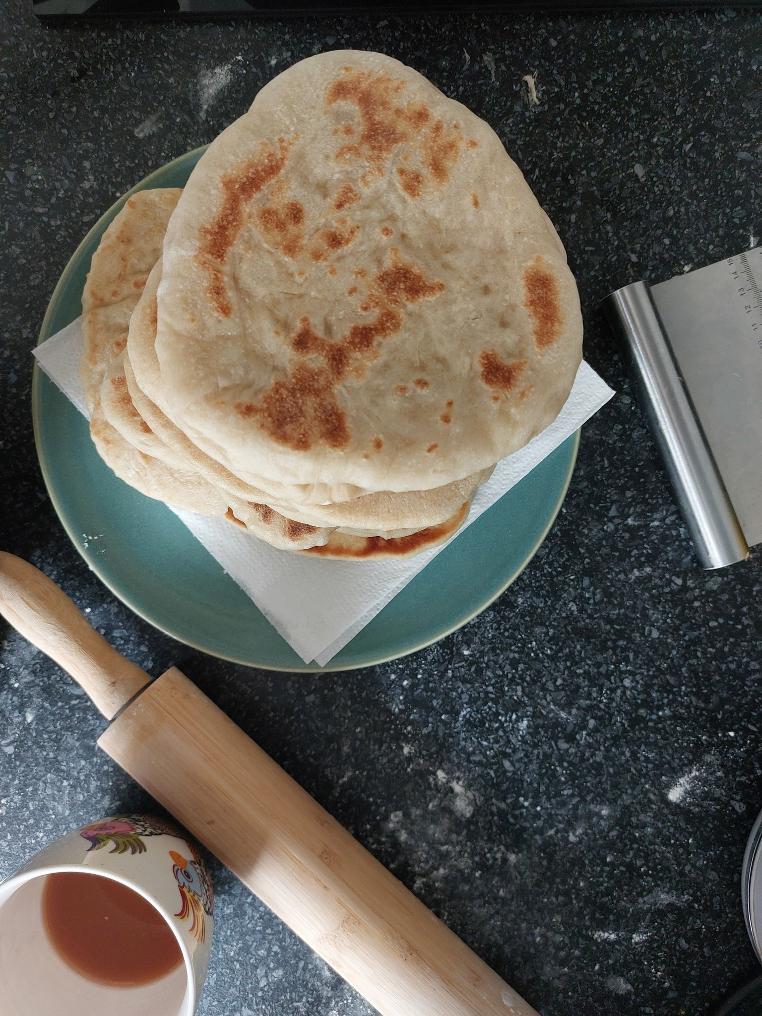 An aerial view of a pile of flatbreads on a turquoise plate with a rolling pin, mug of tea and dough scraper